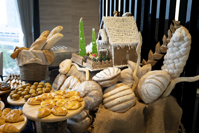 A beautifully arranged display of various types of bread and pastries on a table. There is a gingerbread house decorated with icing, placed on top of several loaves of artisan bread. On the left side, there is a basket with baguettes, and round pastries are neatly placed on wooden stands. The setup includes decorative elements like small Christmas trees and burlap cloth.