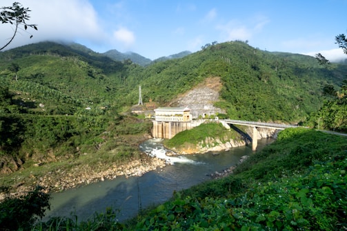 A hydroelectric dam situated in a lush, verdant landscape with dense green hills surrounding it. The structure is built over a river with clear water flowing downstream. The scenery includes a mix of natural greenery and engineered construction, with a bridge connecting across the river adjacent to the dam. The sky is clear with some clouds, conveying a serene atmosphere.