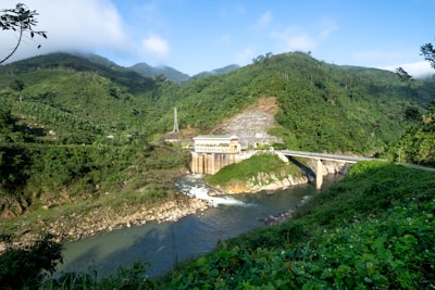 A hydroelectric dam situated in a lush, verdant landscape with dense green hills surrounding it. The structure is built over a river with clear water flowing downstream. The scenery includes a mix of natural greenery and engineered construction, with a bridge connecting across the river adjacent to the dam. The sky is clear with some clouds, conveying a serene atmosphere.