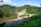 A hydroelectric dam situated in a lush, verdant landscape with dense green hills surrounding it. The structure is built over a river with clear water flowing downstream. The scenery includes a mix of natural greenery and engineered construction, with a bridge connecting across the river adjacent to the dam. The sky is clear with some clouds, conveying a serene atmosphere.