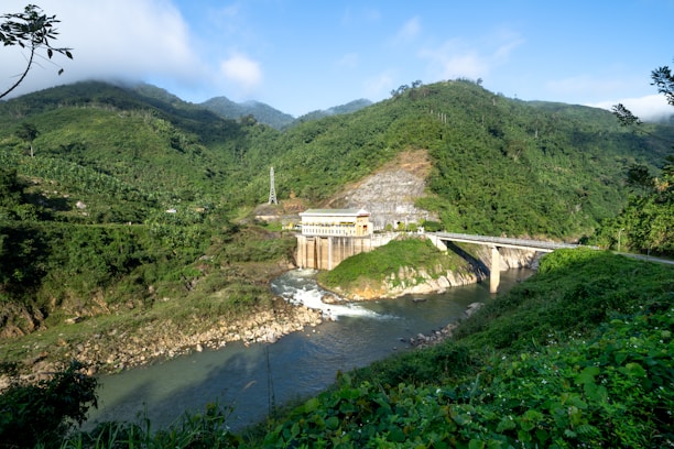 A hydroelectric dam situated in a lush, verdant landscape with dense green hills surrounding it. The structure is built over a river with clear water flowing downstream. The scenery includes a mix of natural greenery and engineered construction, with a bridge connecting across the river adjacent to the dam. The sky is clear with some clouds, conveying a serene atmosphere.