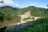 A hydroelectric dam situated in a lush, verdant landscape with dense green hills surrounding it. The structure is built over a river with clear water flowing downstream. The scenery includes a mix of natural greenery and engineered construction, with a bridge connecting across the river adjacent to the dam. The sky is clear with some clouds, conveying a serene atmosphere.