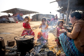 A group of children and an adult are sitting around an outdoor cooking area with a pot on fire. The children are wearing colorful traditional clothing. A motorcycle is parked in the background near some makeshift shelters. The atmosphere is rustic and informal.
