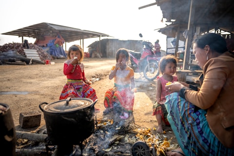 A group of children and an adult are sitting around an outdoor cooking area with a pot on fire. The children are wearing colorful traditional clothing. A motorcycle is parked in the background near some makeshift shelters. The atmosphere is rustic and informal.