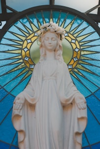 A serene marble statue of a woman with a floral crown, positioned in front of a decorative stained glass backdrop featuring vibrant blue and yellow patterns. The figure is draped in a robe and holds her arms slightly outstretched.