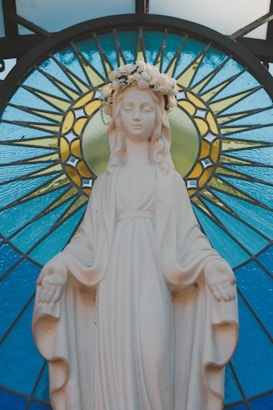 A serene marble statue of a woman with a floral crown, positioned in front of a decorative stained glass backdrop featuring vibrant blue and yellow patterns. The figure is draped in a robe and holds her arms slightly outstretched.
