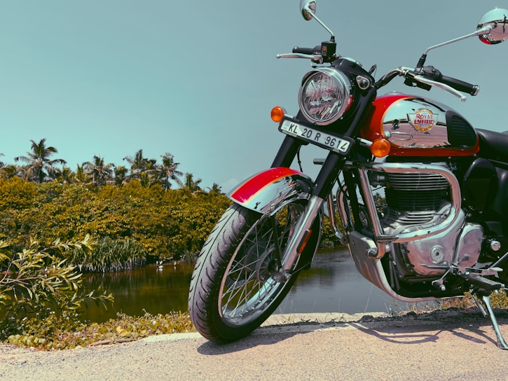 A Royal Enfield motorcycle is parked on a road next to a scenic body of water. The motorcycle has a bright red accent with shiny chrome details. Behind it, there are lush green trees and a clear blue sky.