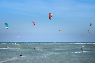 Multiple kite surfers are gliding across the ocean waters, each controlling a colorful kite that soars high above. The sky is clear with a soft blue color, and the ocean looks dynamic with visible waves.