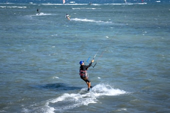 A person is engaged in kitesurfing on a vast, open sea. The individual is wearing a wetsuit and a blue cap, gripping the kite handle firmly while gliding over the water. Other kitesurfers can be seen in the background, adding to the dynamic and lively scene.