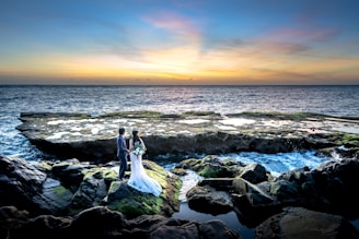 A couple stands on rocky outcrops by the ocean, with the woman in a wedding dress holding a bouquet and the man in a suit. The sunset creates a colorful gradient in the sky, reflecting on the water. Waves crash gently against the rocks, which are covered in patches of green moss.