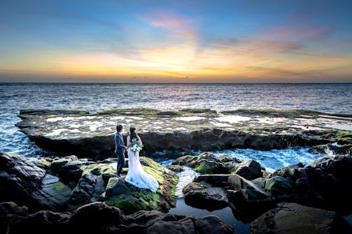 A couple stands on rocky outcrops by the ocean, with the woman in a wedding dress holding a bouquet and the man in a suit. The sunset creates a colorful gradient in the sky, reflecting on the water. Waves crash gently against the rocks, which are covered in patches of green moss.