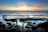 A couple stands on rocky outcrops by the ocean, with the woman in a wedding dress holding a bouquet and the man in a suit. The sunset creates a colorful gradient in the sky, reflecting on the water. Waves crash gently against the rocks, which are covered in patches of green moss.