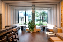 A modern interior featuring a spacious room with wooden flooring and wooden seating areas. The ceiling and walls are adorned with geometric frames that create an artistic ceiling design. Large windows allow for ample natural light, revealing a view of outdoor greenery. Green plants are strategically placed inside the room for a touch of nature.