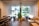 A modern interior featuring a spacious room with wooden flooring and wooden seating areas. The ceiling and walls are adorned with geometric frames that create an artistic ceiling design. Large windows allow for ample natural light, revealing a view of outdoor greenery. Green plants are strategically placed inside the room for a touch of nature.