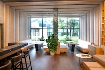 A modern interior featuring a spacious room with wooden flooring and wooden seating areas. The ceiling and walls are adorned with geometric frames that create an artistic ceiling design. Large windows allow for ample natural light, revealing a view of outdoor greenery. Green plants are strategically placed inside the room for a touch of nature.
