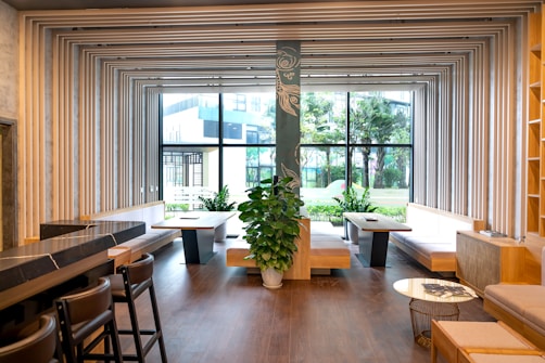 A modern interior featuring a spacious room with wooden flooring and wooden seating areas. The ceiling and walls are adorned with geometric frames that create an artistic ceiling design. Large windows allow for ample natural light, revealing a view of outdoor greenery. Green plants are strategically placed inside the room for a touch of nature.