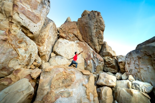 A person wearing a red shirt is climbing amidst large, rugged rock formations under a clear blue sky. The rocks vary in color from tan to brown and are weathered with unique patterns.
