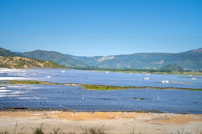 A vast solar panel farm is spread across a flat landscape with clear blue skies and distant green hills in the background. The panels are organized in neat rows, reflecting sunlight.