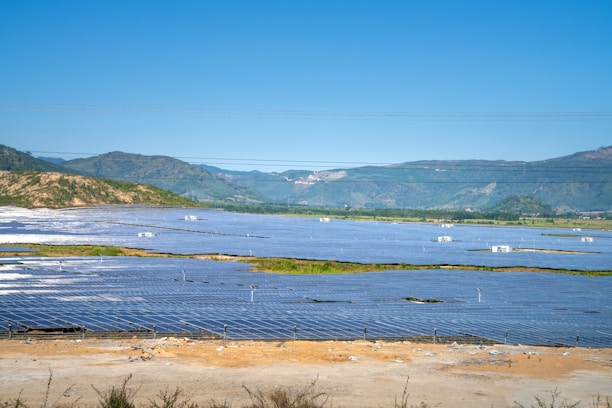 A vast solar panel farm is spread across a flat landscape with clear blue skies and distant green hills in the background. The panels are organized in neat rows, reflecting sunlight.