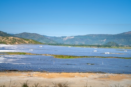 A vast solar panel farm is spread across a flat landscape with clear blue skies and distant green hills in the background. The panels are organized in neat rows, reflecting sunlight.