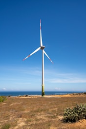 A large wind turbine with three blades stands prominently on a grassy landscape under a clear blue sky. In the background, the ocean is visible, contributing to the serene setting. The turbine’s blades have red markings at the tips, and the structure's base is green.