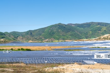 Expansive solar panel installation spread across a large, open area, nestled in front of lush green hills under a clear blue sky.