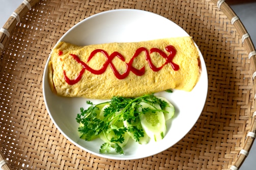 A neatly crafted omelette is adorned with a zigzag pattern of red ketchup, resting on a white plate. Fresh green cilantro and slices of cucumber accompany the omelette on the plate. The setting includes a woven wooden mat that provides a rustic backdrop.