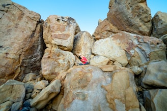 A climber wearing a red shirt scales a large, rugged rock face bordered by enormous boulders under a clear blue sky.