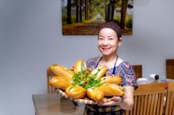 A woman stands indoors holding a large tray of filled baguettes garnished with herbs. She is smiling and wearing a printed dress with a headband. Behind her, there's a dining area with wooden chairs and a wall-mounted piece of art depicting a scenic forest path.