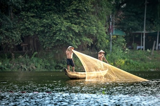 Two individuals wearing traditional conical hats are fishing from a wooden boat on a calm river, surrounded by dense greenery. They are casting a large, golden fishing net into the water, creating a graceful arc.