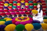 A vibrant display of colorful incense sticks arranged in layers and patterns, with shades of red, yellow, green, blue, and purple. A woman dressed in a traditional white outfit kneels beside the incense sticks, holding a bunch of yellow ones.