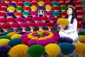 A vibrant display of colorful incense sticks arranged in layers and patterns, with shades of red, yellow, green, blue, and purple. A woman dressed in a traditional white outfit kneels beside the incense sticks, holding a bunch of yellow ones.