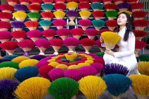 A vibrant display of colorful incense sticks arranged in layers and patterns, with shades of red, yellow, green, blue, and purple. A woman dressed in a traditional white outfit kneels beside the incense sticks, holding a bunch of yellow ones.