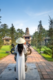 A person wearing a traditional conical hat and long white dress stands in front of an ornate stone sculpture. The scene is set in a garden with a paved path leading towards a historical tower surrounded by trees.