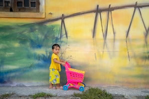 A young child wearing a yellow shirt and floral pants holds a pink toy shopping cart in front of a vibrant, painted wall. The wall features an abstract landscape with shades of green, blue, and yellow, including elements resembling a fence and a window in the upper left corner.