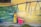A young child wearing a yellow shirt and floral pants holds a pink toy shopping cart in front of a vibrant, painted wall. The wall features an abstract landscape with shades of green, blue, and yellow, including elements resembling a fence and a window in the upper left corner.