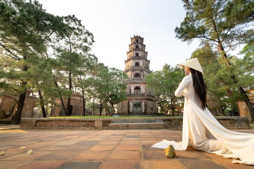 A woman wearing a traditional conical hat and a long white dress sits on a tiled path, looking towards a historical multi-tiered pagoda surrounded by lush trees.