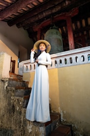 A woman wearing a traditional white dress and a conical hat stands on an old stone staircase. She is positioned under a weathered wooden structure, with a large bell behind her. The setting appears to be an ancient or historic site.