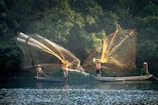 Fishermen are casting large nets from small wooden boats on a calm river, surrounded by dense green foliage. The sunlight illuminates the golden nets and creates reflections on the water.