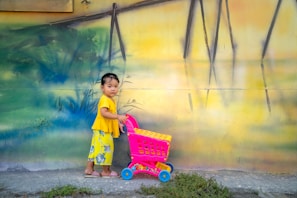 A young child wearing a bright yellow outfit stands next to a vibrant pink toy shopping cart in front of a colorful mural. The wall is adorned with abstract patterns, combining shades of green, blue, and yellow. The child appears to be calmly looking at the camera.