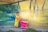 A young child wearing a bright yellow outfit stands next to a vibrant pink toy shopping cart in front of a colorful mural. The wall is adorned with abstract patterns, combining shades of green, blue, and yellow. The child appears to be calmly looking at the camera.