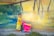 A young child wearing a bright yellow outfit stands next to a vibrant pink toy shopping cart in front of a colorful mural. The wall is adorned with abstract patterns, combining shades of green, blue, and yellow. The child appears to be calmly looking at the camera.