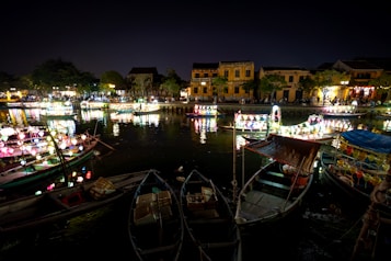 A picturesque riverside scene at night with traditional boats adorned with colorful lanterns reflecting on the water. The ambiance is lit by warm, vibrant lights from the lanterns and nearby buildings with classic architectural designs. The street by the riverbank is populated with people enjoying the view.