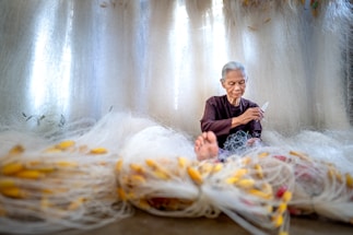 An elderly person with gray hair is seated among an array of large fishing nets, intricately working on them with focused concentration. The nets are spread out around the individual, featuring a multitude of bright yellow floats attached. The background is filled with more nets, creating a softly lit, serene atmosphere. The person wears a dark, simple garment and appears to be skilled at their task.