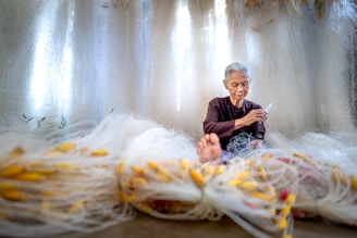 An elderly person with gray hair is seated among an array of large fishing nets, intricately working on them with focused concentration. The nets are spread out around the individual, featuring a multitude of bright yellow floats attached. The background is filled with more nets, creating a softly lit, serene atmosphere. The person wears a dark, simple garment and appears to be skilled at their task.
