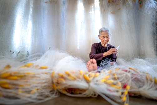 An elderly person with gray hair is seated among an array of large fishing nets, intricately working on them with focused concentration. The nets are spread out around the individual, featuring a multitude of bright yellow floats attached. The background is filled with more nets, creating a softly lit, serene atmosphere. The person wears a dark, simple garment and appears to be skilled at their task.