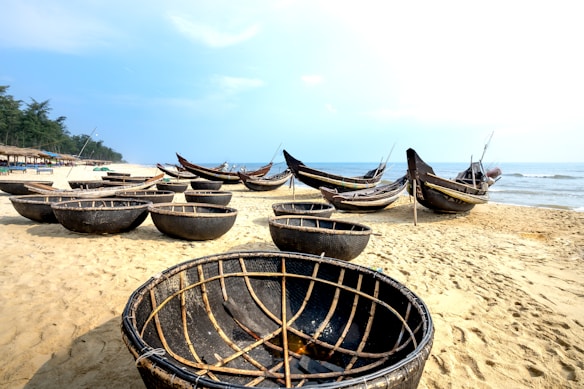 A sandy beach scene with traditional round basket boats and long wooden fishing boats resting on the shore. The horizon shows a calm sea meeting a blue sky. In the background, there are rows of umbrellas and a line of trees, indicating a beach resort area.