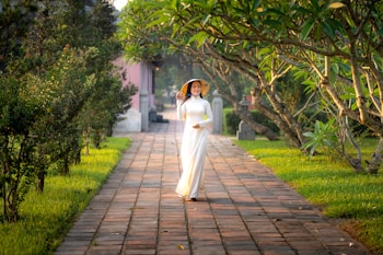 A woman wearing a traditional white áo dài dress and a conical hat walks down a paved pathway surrounded by lush greenery and trees. The scene is serene and calm, with sunlight filtering through the leaves, creating dappled shadows on the path. The atmosphere suggests a peaceful garden or park setting.