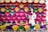 A woman in a white traditional outfit is standing amidst an array of colorful incense sticks arranged in organized patterns. The sticks are fanned out, displaying vibrant colors such as red, pink, purple, yellow, and green. The backdrop also includes a collection of conical hats at the top.