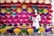 A woman in a white traditional outfit is standing amidst an array of colorful incense sticks arranged in organized patterns. The sticks are fanned out, displaying vibrant colors such as red, pink, purple, yellow, and green. The backdrop also includes a collection of conical hats at the top.